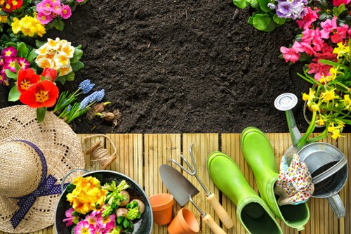 The image displays an organized garden scene from a high angle, featuring a rectangular area with dark, freshly turned soil bordered by colorful flowering plants, including pink, yellow, and purple blossoms, along the top edge. In the foreground, there is a wooden deck or patio with various gardening tools and accessories, such as a pair of bright green rubber boots, a watering can filled with small multicoloured flowers, a trowel, a garden fork, and a small spade. A straw sunhat with a purple ribbon rests on the corner of the deck, adding a touch of outdoor leisure. The garden shows a well-maintained lawn or grassy area in the background, and the scene gives an impression of ongoing gardening activities typical for a backyard or front garden in Willesden, with natural lighting suggesting a sunny day. This setting aligns with gardening services focused on lawn and plant care, landscaping, and outdoor maintenance, highlighting elements common to residential gardens in the area. The image provides a clear, detailed view of garden organization and tools used for outdoor work, emphasizing a tidy and inviting outdoor space.