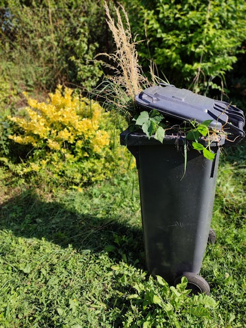 Two green wheelie bins branded with 'SUEZ' are positioned on the edge of a paved sidewalk, nearby a small dirt patch with some scattered leaves and debris. Behind the bins, a street with an asphalt su