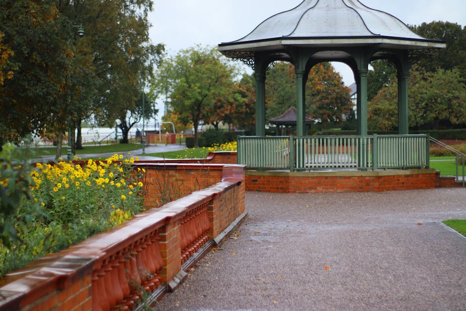 A landscaped park area featuring a circular gazebo with a metal roof supported by decorative columns, surrounded by a low brick wall with a green wooden railing. The foreground shows a curved brick fl